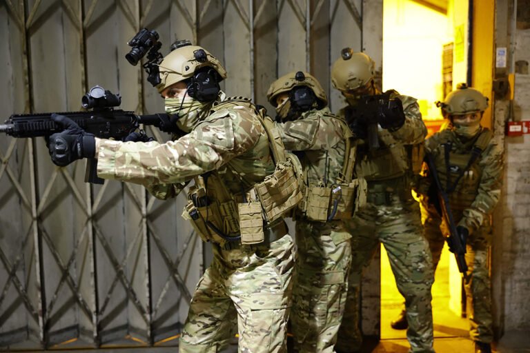 Four soldiers are scanning an abandoned building. They are wearing military camo trousers and tops, ballistic helmets, ballistic plate carriers, gloves and guns.